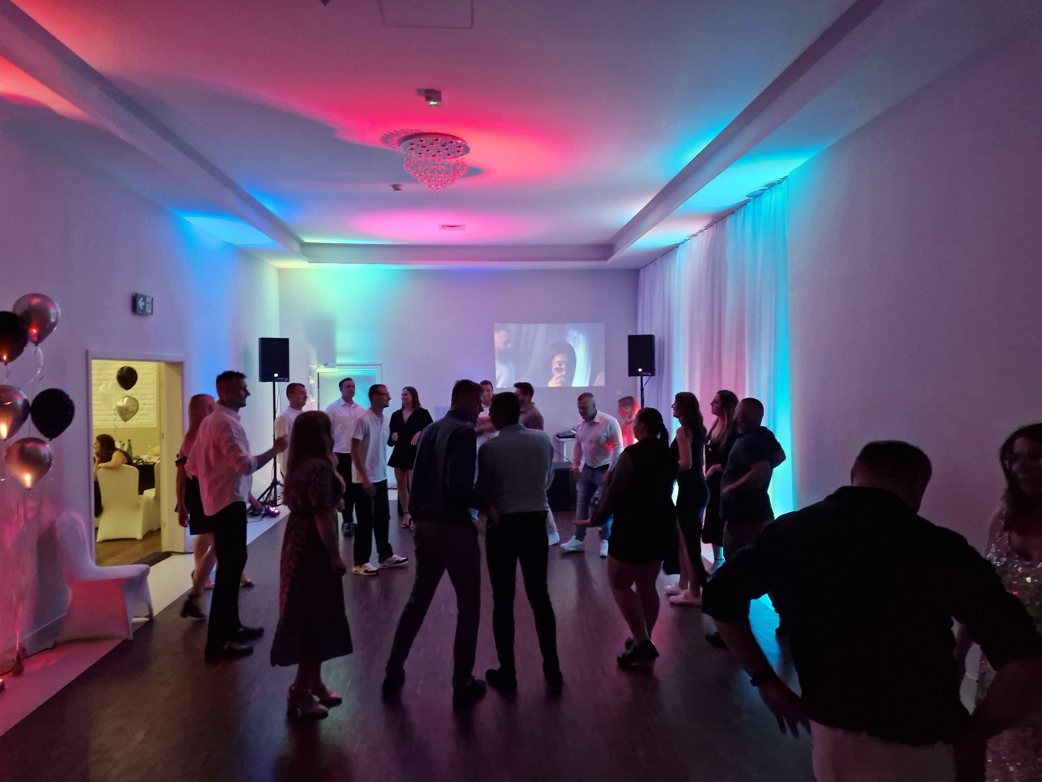 Impreza firmowa w sali z oświetleniem LED: tańczący ludzie, balony, projektor wyświetlający obraz. Kolorowe światła na suficie i ścianach tworzą radosną atmosferę.