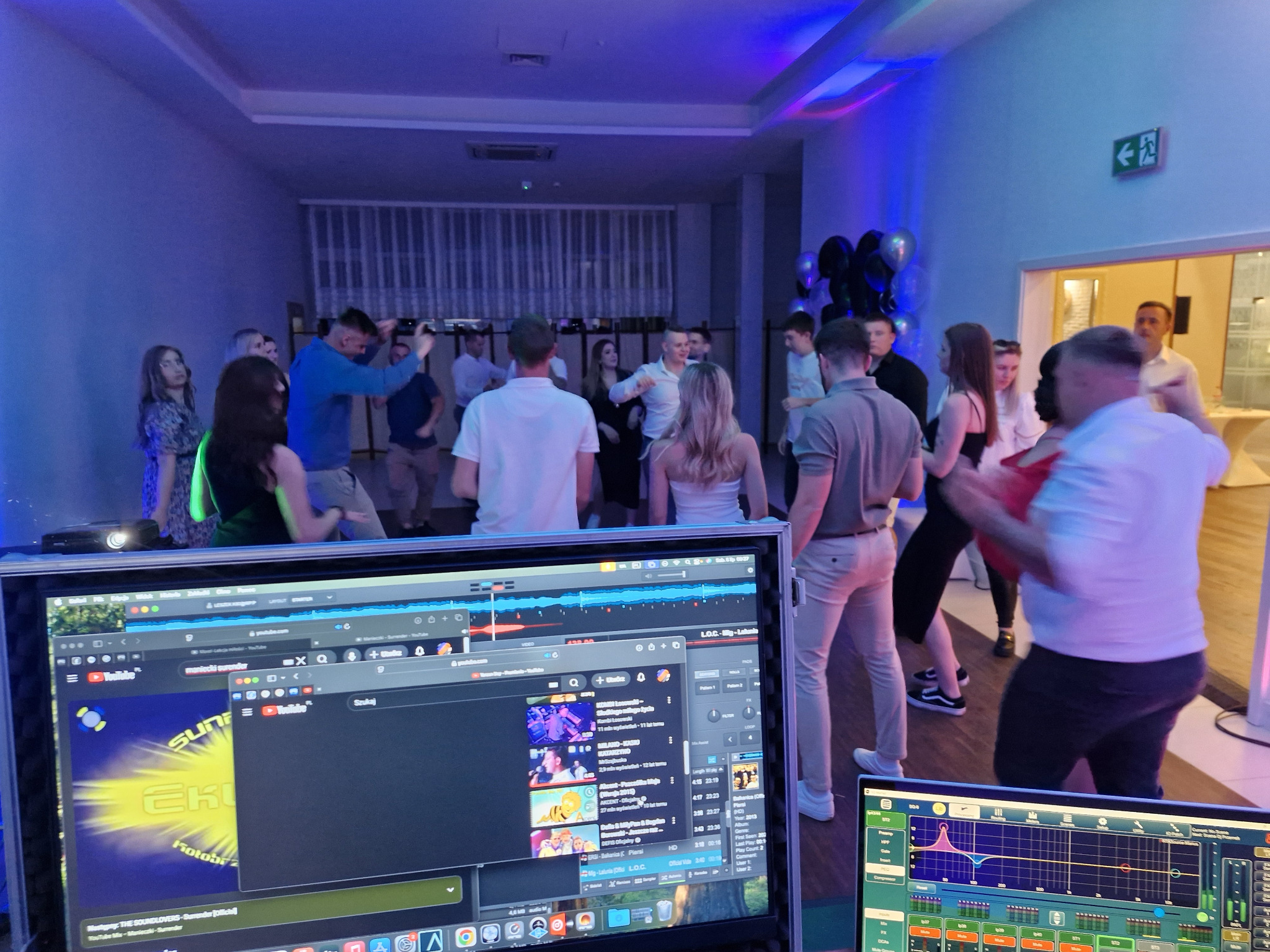 Impreza firmowa w niebieskim oświetleniu, z widocznymi monitorami DJ-a i tańczącymi osobami w tle. Dynamiczna atmosfera zabawy.