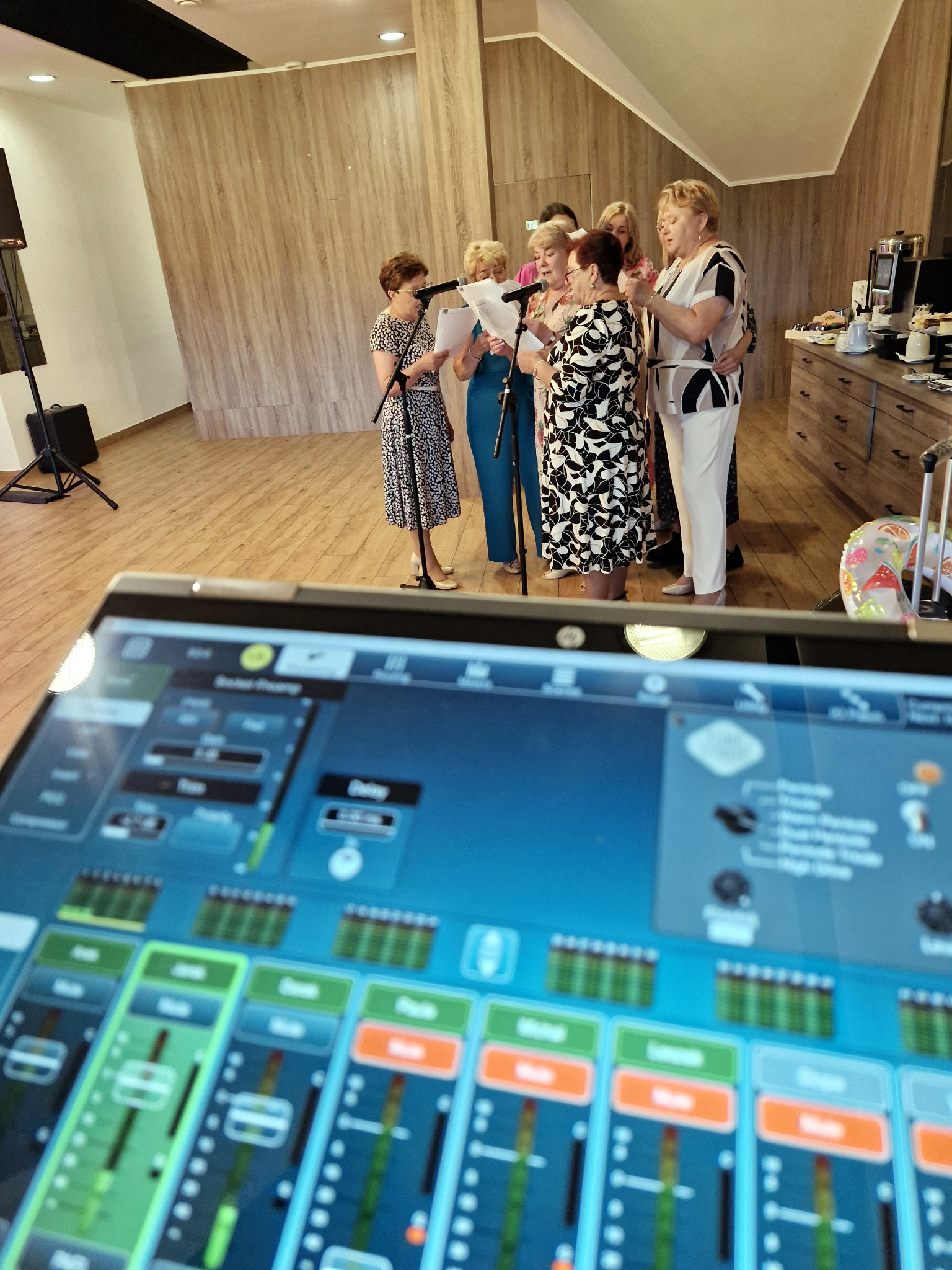Grupa kobiet śpiewa do mikrofonów, widok zza konsolety mikserskiej. Jasne wnętrze z drewnianymi panelami. Sprzęt nagłośnieniowy w tle.
