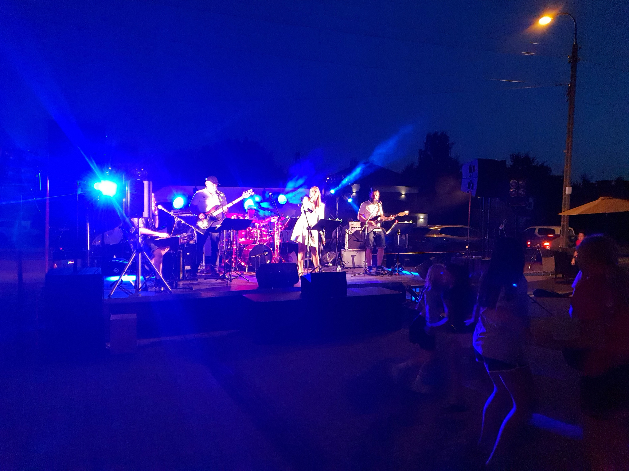 Zespół muzyczny na scenie plenerowej, oświetlony niebieskimi światłami, w tle publiczność. Wokalistka w białej sukience, gitarzyści i perkusista. Wieczorny koncert.