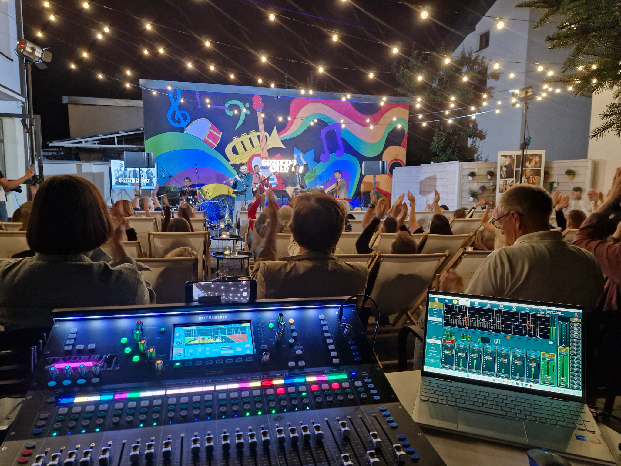 Koncert plenerowy z zespołem na scenie, widok zza konsolety mikserskiej i laptopa z oprogramowaniem audio, publiczność w tle. Oświetlenie sceniczne i dekoracyjne.