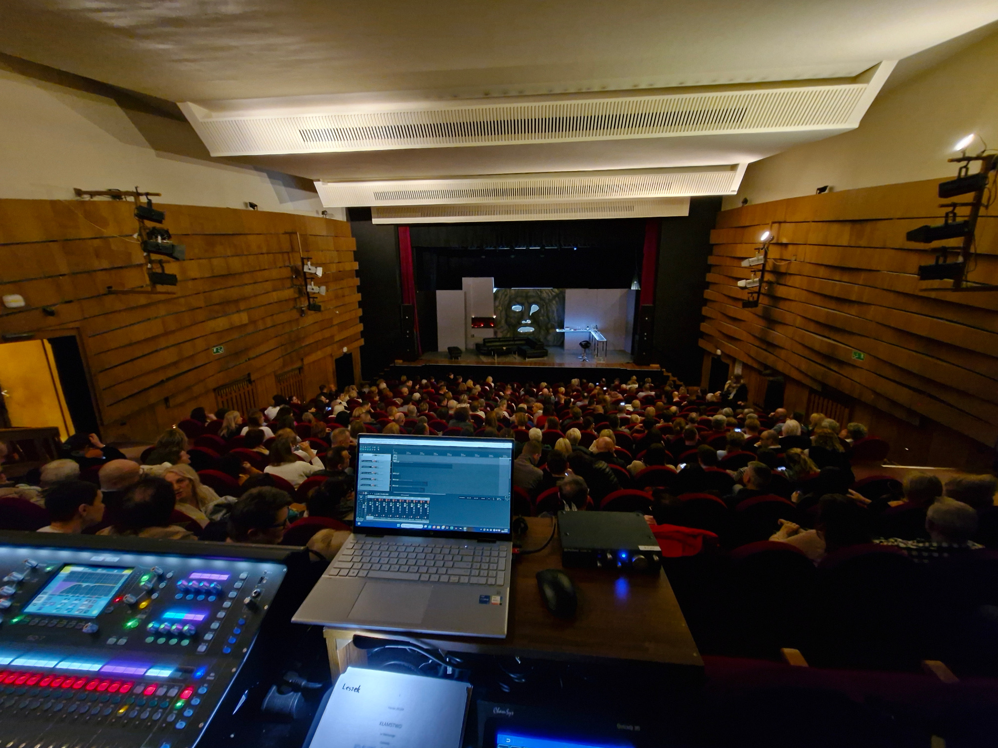 Sala teatralna wypełniona publicznością, widok zza konsolety dźwiękowej z laptopem, oświetlenie sceniczne. Przygotowania do spektaklu.
