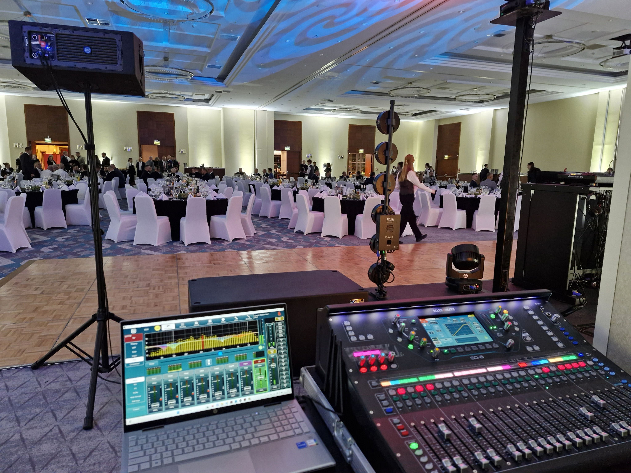 Sala bankietowa z ustawionym sprzętem nagłośnieniowym: mikser, laptop z analizatorem dźwięku, głośnik na statywie. W tle stoliki z gośćmi.