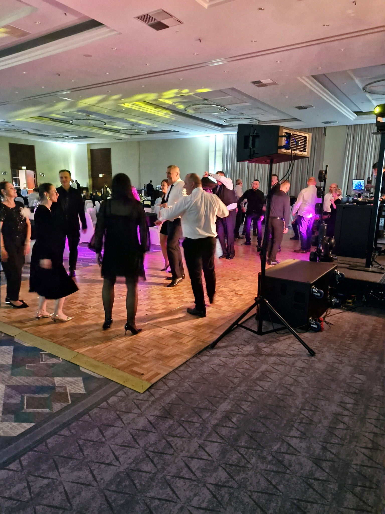 Parkiet taneczny wypełniony tańczącymi osobami w strojach wieczorowych, z widocznym sprzętem nagłośnieniowym i oświetleniem w tle sali balowej.