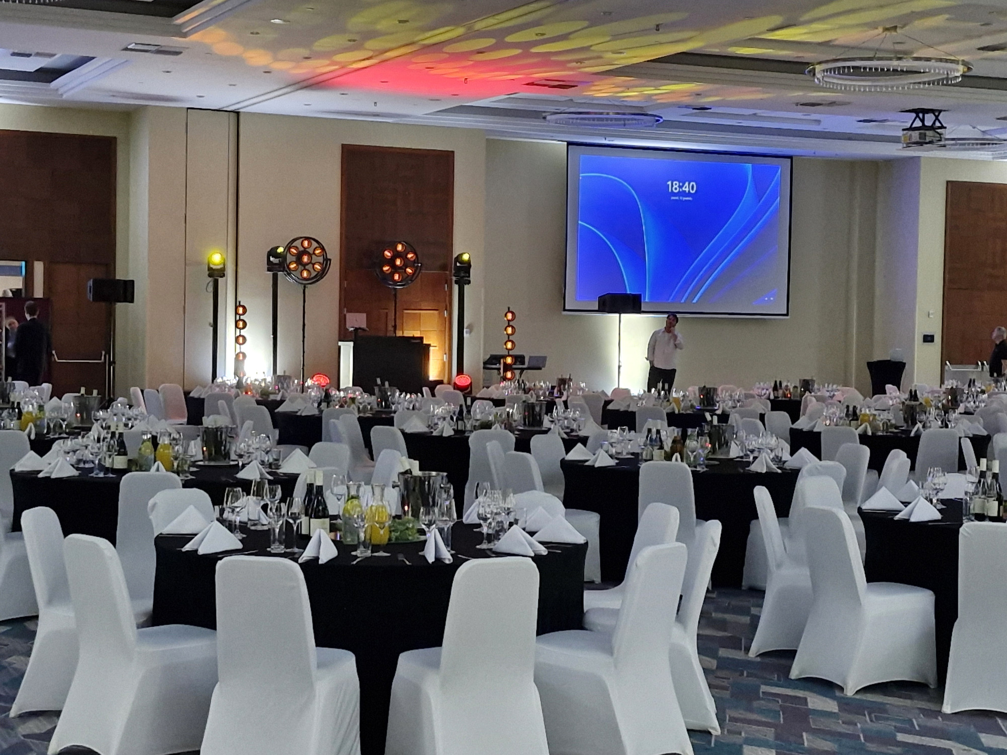 Sala bankietowa z oświetleniem eventowym, stołami nakrytymi do uroczystości i ekranem projekcyjnym. Przygotowania do wydarzenia.