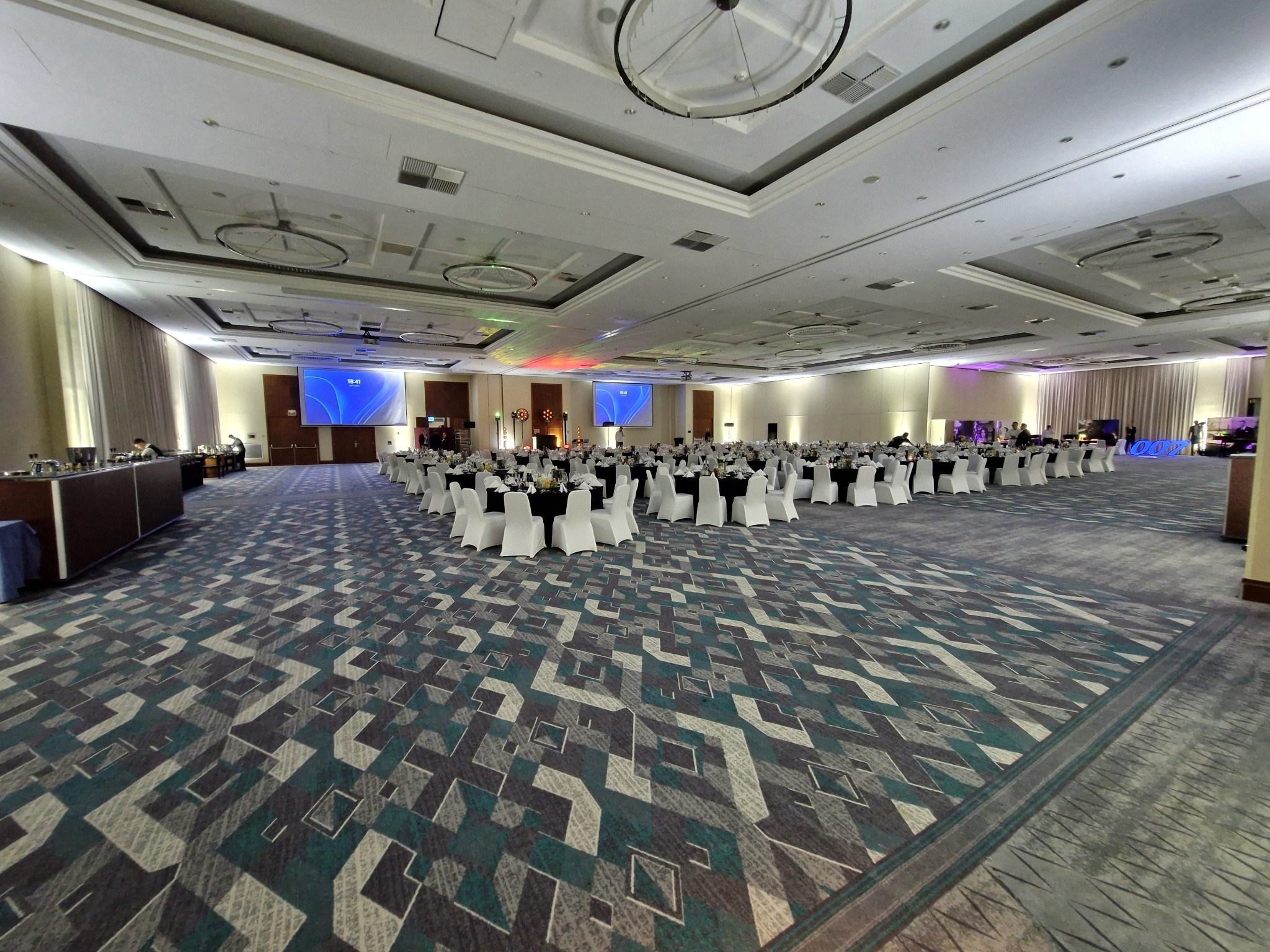 Sala bankietowa z okrągłymi stołami, białymi krzesłami i dekoracjami świetlnymi. Duża przestrzeń gotowa na imprezę, widok z perspektywy szerokokątnej.