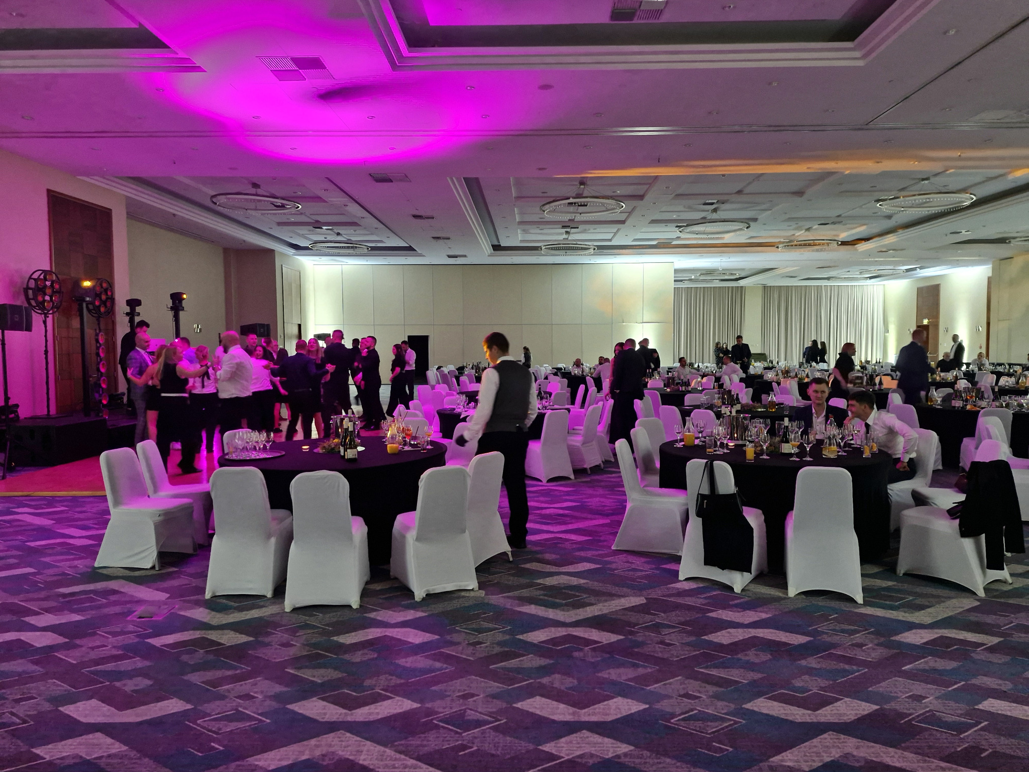 Sala bankietowa z okrągłymi stołami, białymi krzesłami i fioletowym oświetleniem. Goście w tle, przygotowania do wydarzenia, parkiet z wzorem.