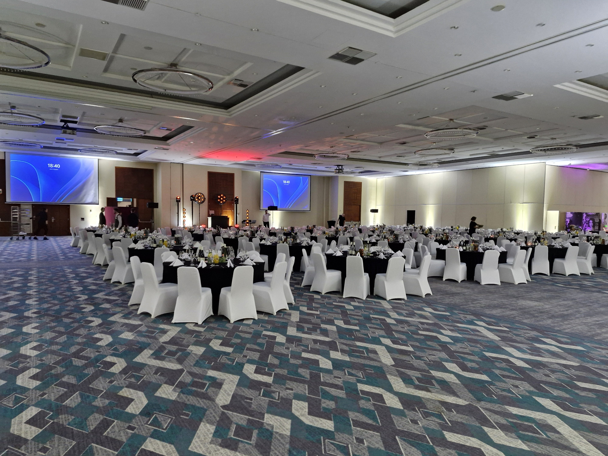 Sala bankietowa z ustawionymi stołami i krzesłami, gotowa na wydarzenie. Widoczne ekrany i oświetlenie sceniczne. Wzór dywanu w odcieniach szarości i błękitu.