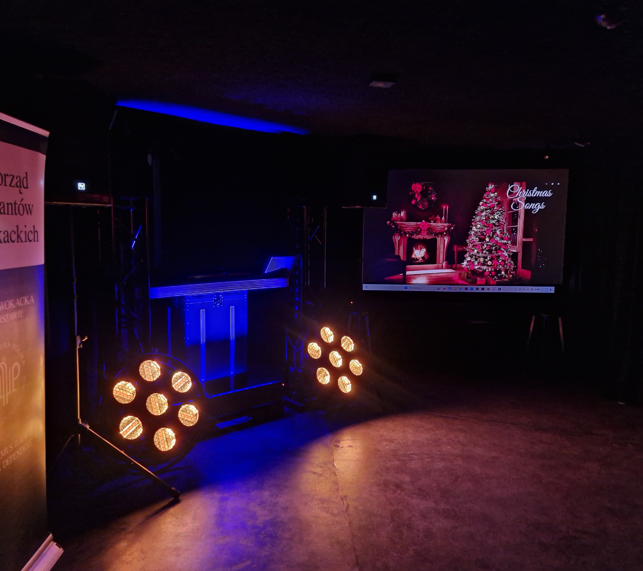 Scena z ekranem wyświetlającym świąteczne piosenki, oświetlona reflektorami i niebieskim światłem. Widoczny sprzęt nagłośnieniowy i baner reklamowy z boku.