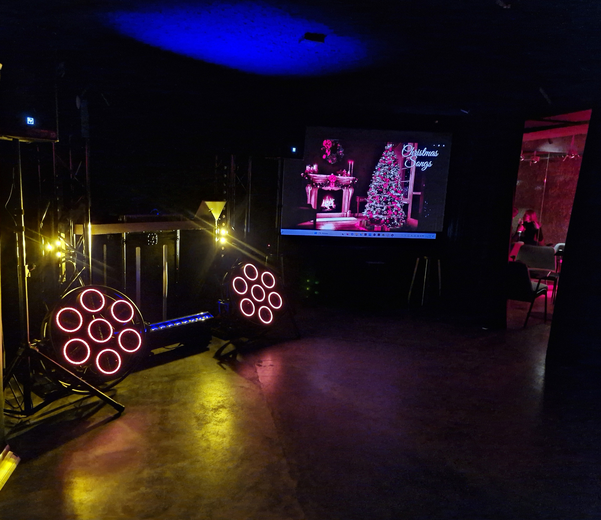 Scena z oświetleniem LED i ekranem z motywem świątecznym. Dwa reflektory z okrągłymi lampami, w tle ekran z choinką i kominkiem. Atmosfera ciemna, klubowa.