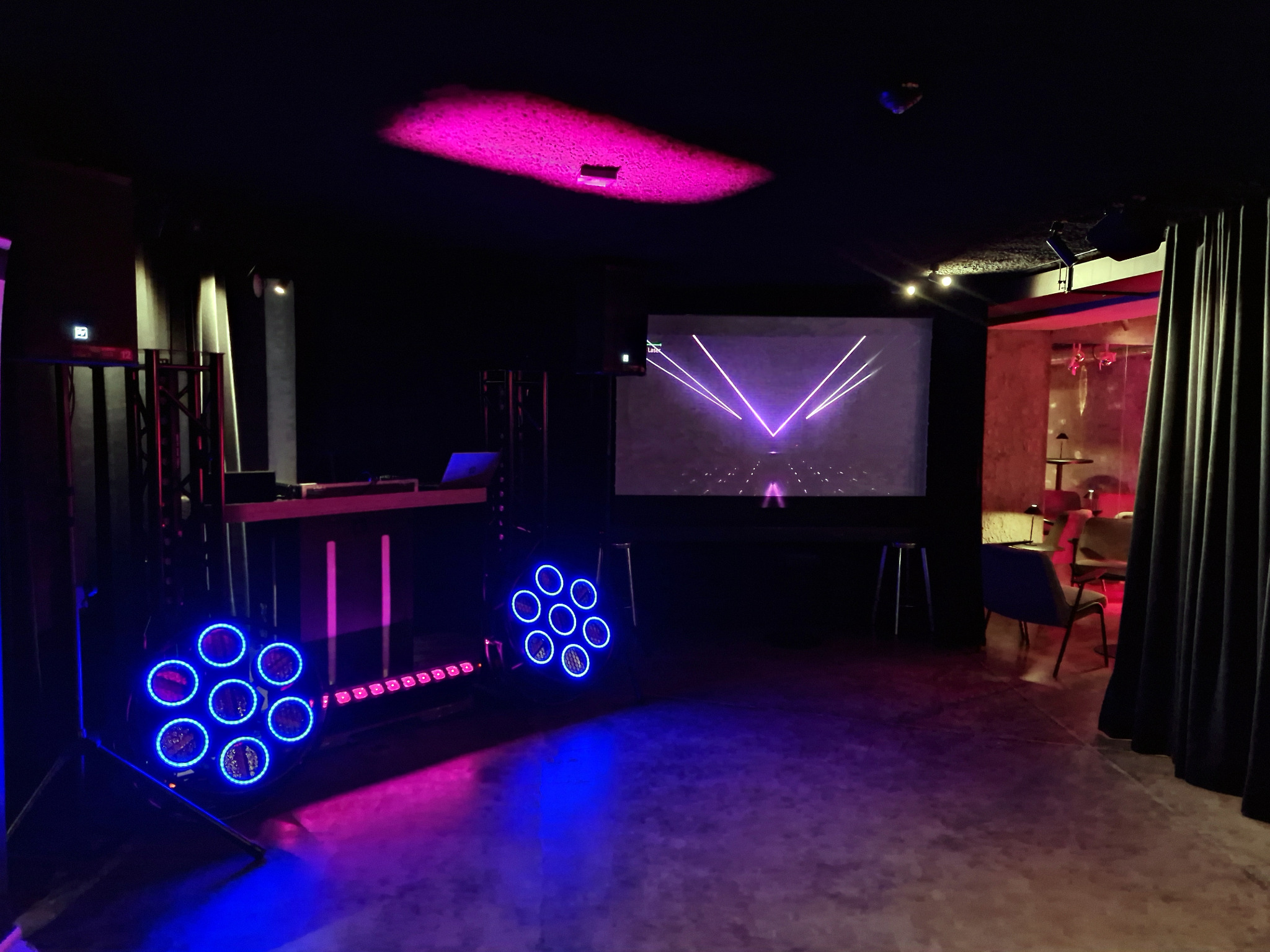 Oświetlenie sceniczne z DJ-ką i ekranem w tle, tworzące klubową atmosferę. Reflektory w kolorze niebieskim i różowym oświetlają parkiet. Widoczny sprzęt nagłośnieniowy.
