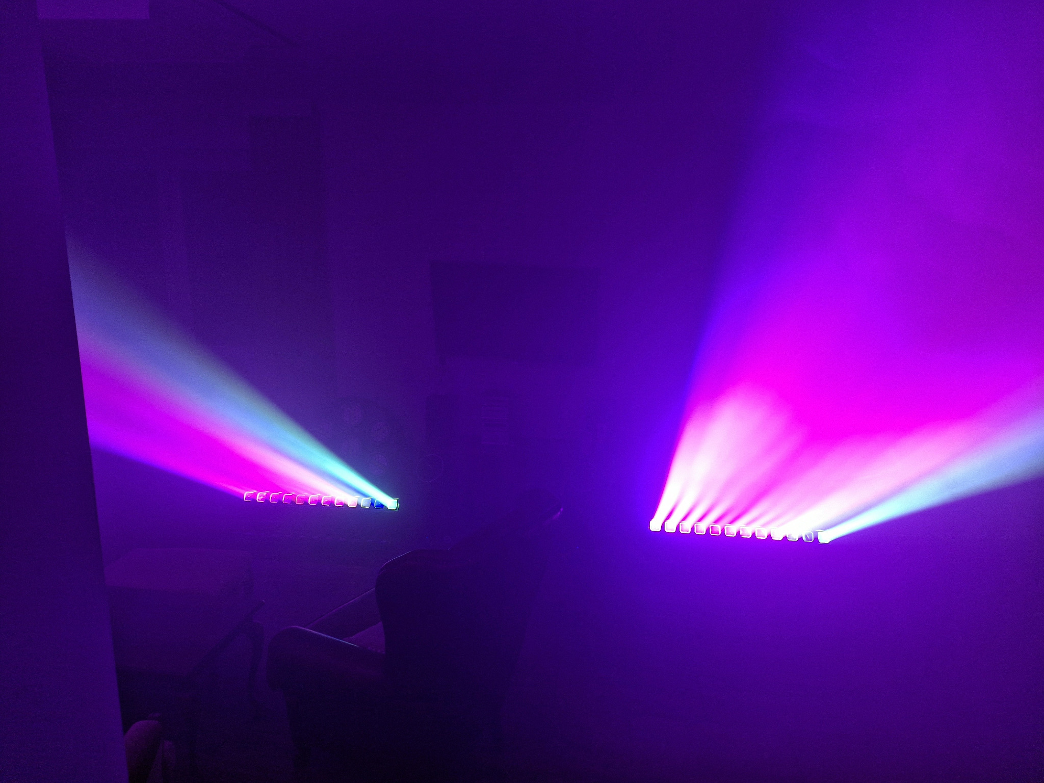 Fioletowe oświetlenie sceniczne z dwoma listwami LED, emitującymi kolorowe promienie światła w zadymionym pomieszczeniu. Widoczne zarysy krzesła i osoby.