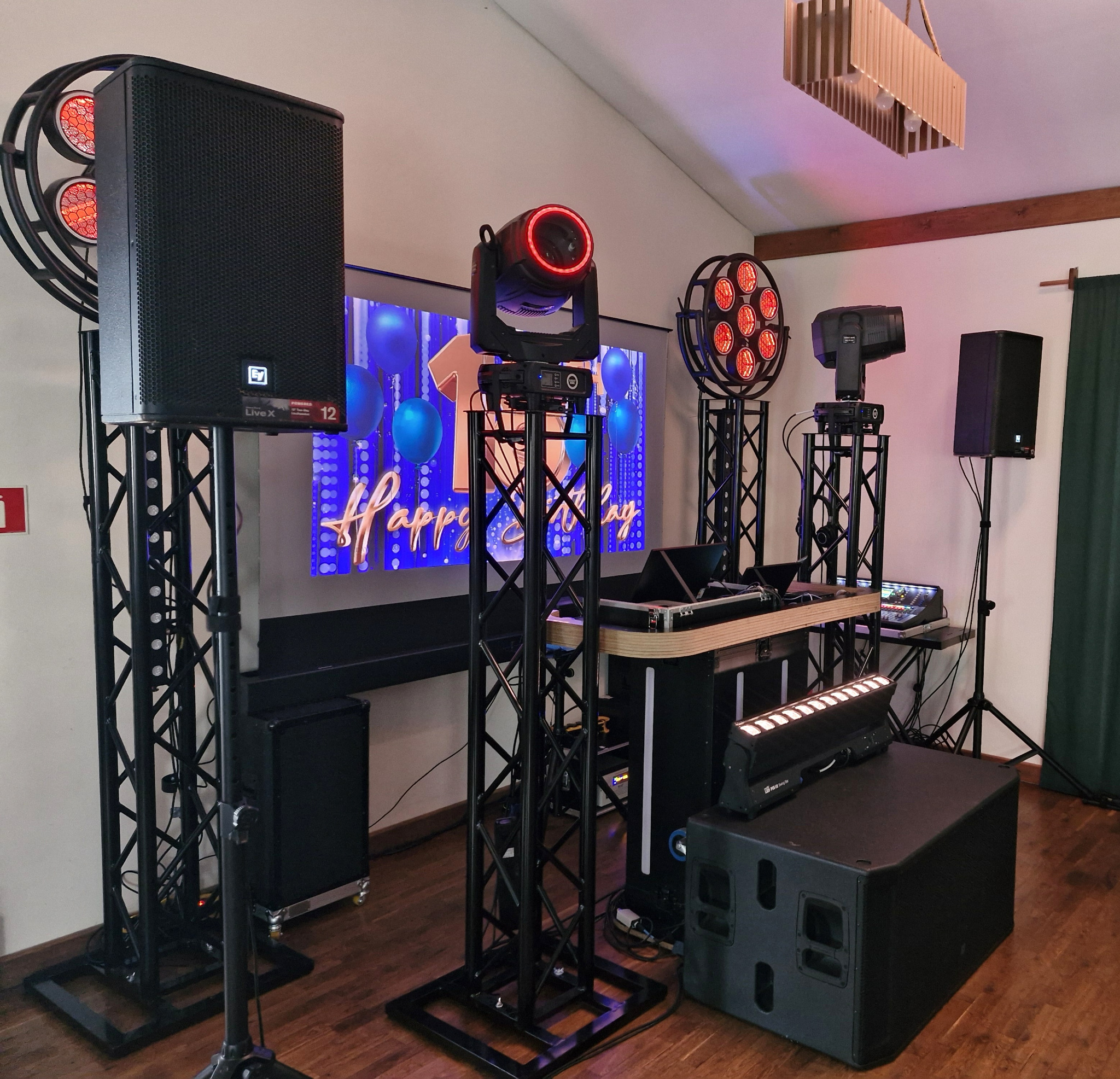 Profesjonalne nagłośnienie i oświetlenie eventowe: kolumny głośnikowe, reflektory sceniczne na statywach, mikser DJ-ski. Tło stanowi dekoracja urodzinowa.