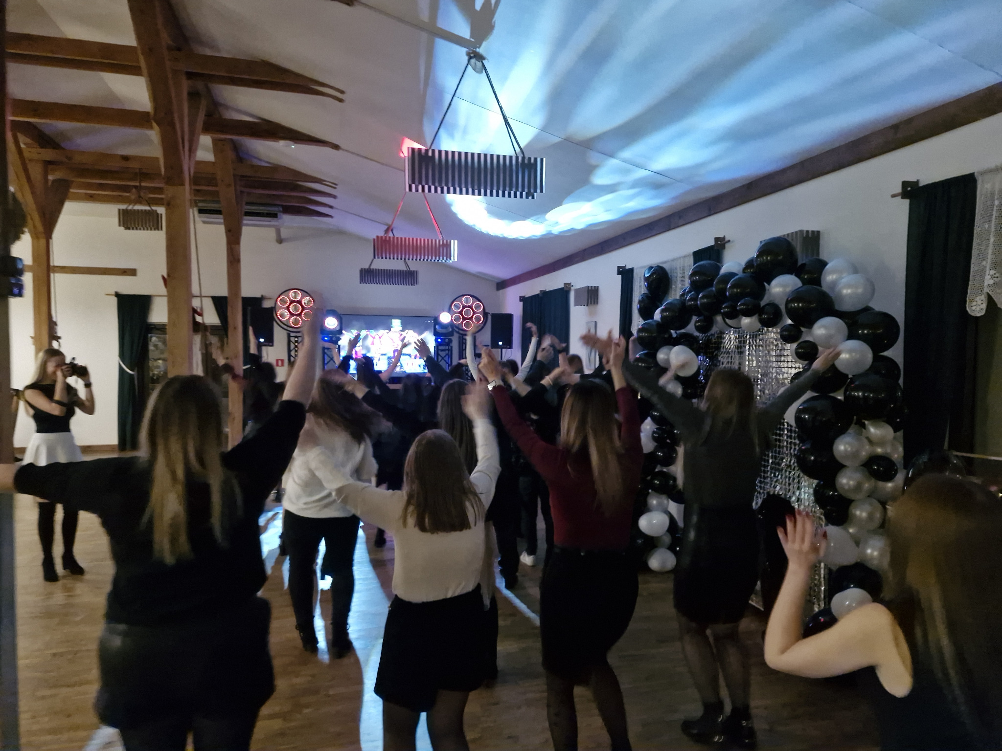 Sala balowa z drewnianymi belkami, oświetleniem scenicznym i dekoracjami z balonów. W tle tańczące osoby, tworząc radosną atmosferę imprezy.