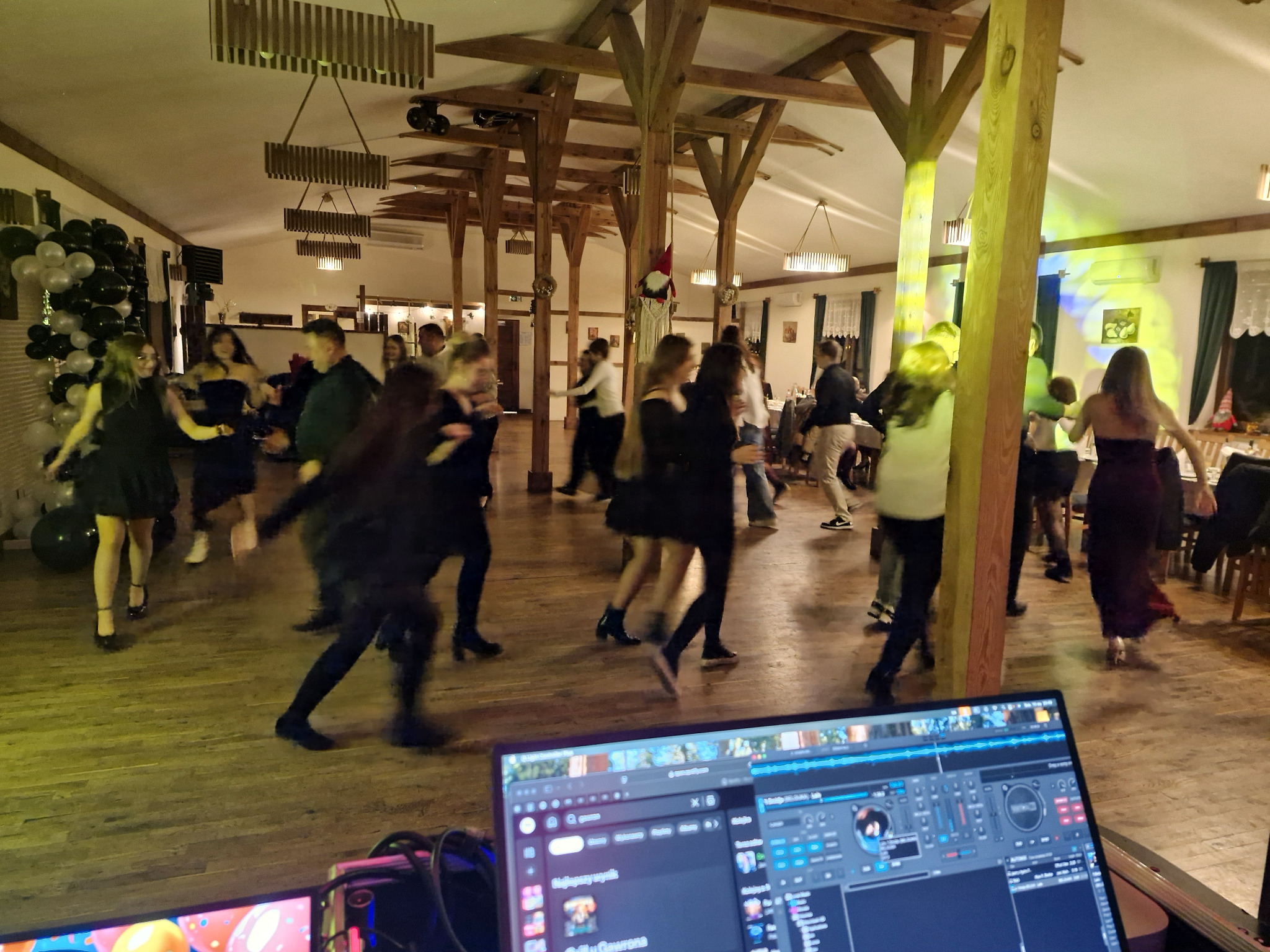 Parkiet taneczny wypełniony bawiącymi się ludźmi, widok zza laptopa DJ-a, drewniana sala balowa z kolumnami i dekoracjami.