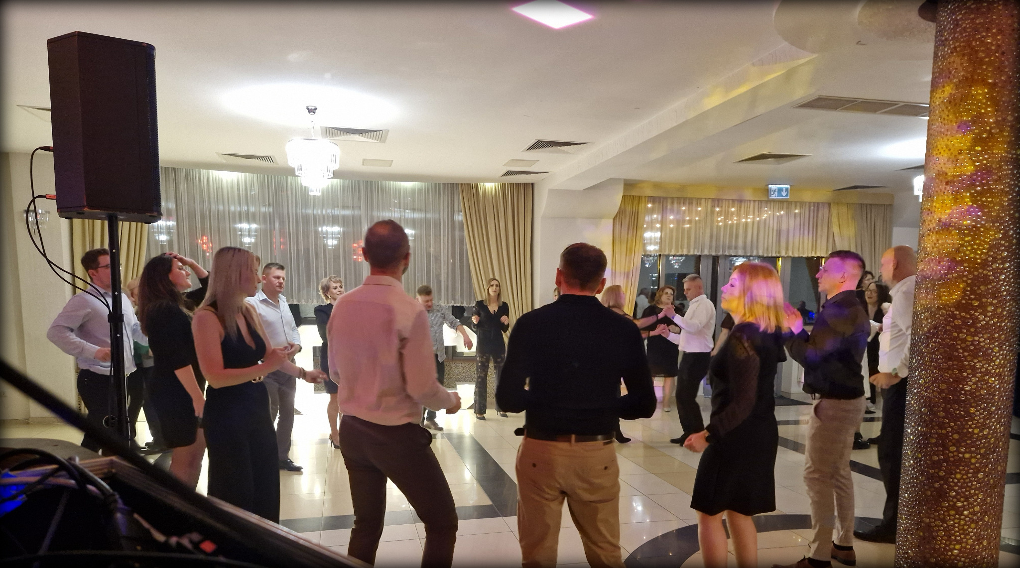 Parkiet taneczny wypełniony gośćmi podczas imprezy w sali balowej z kolumną dekoracyjną i nagłośnieniem. Widok zza konsoli DJ-a.