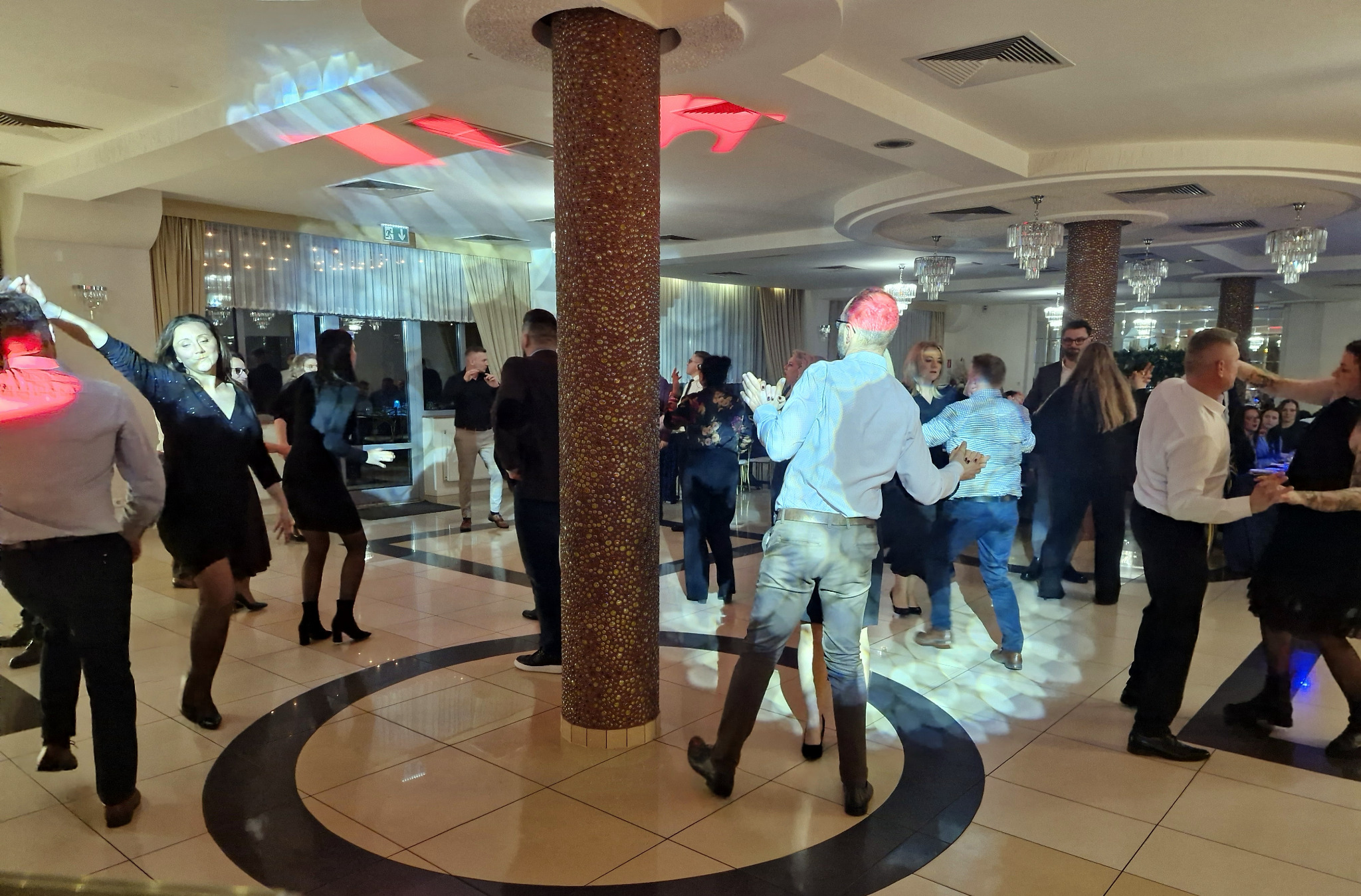 Parkiet taneczny w sali balowej z tańczącymi osobami, kolumna dekoracyjna na środku, oświetlenie sceniczne i eleganckie żyrandole.