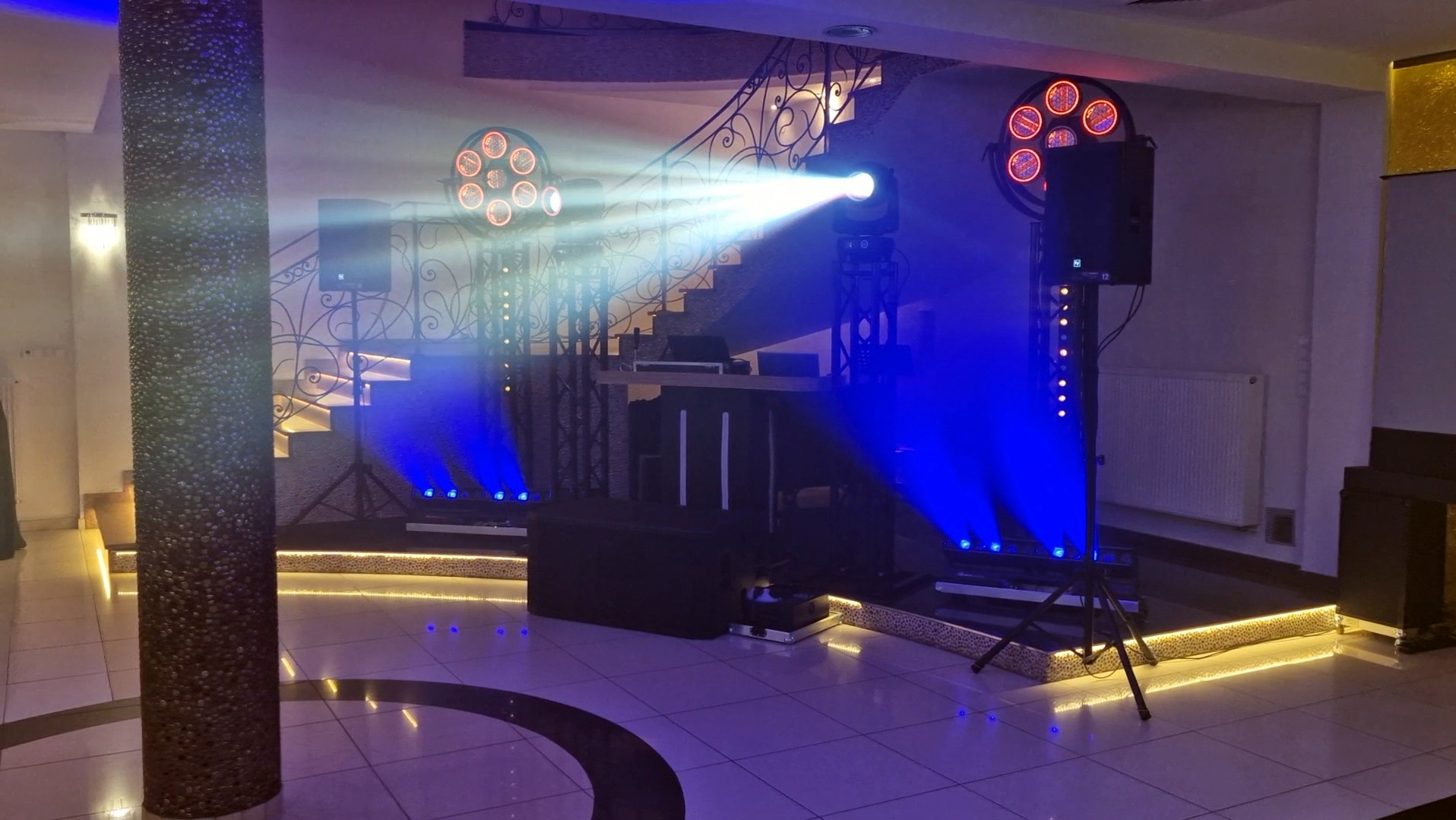 Oświetlenie eventowe z reflektorami i kolumnami głośnikowymi na sali z dekoracyjnymi schodami i kolumną. Niebieskie światło tworzy efektowną atmosferę.