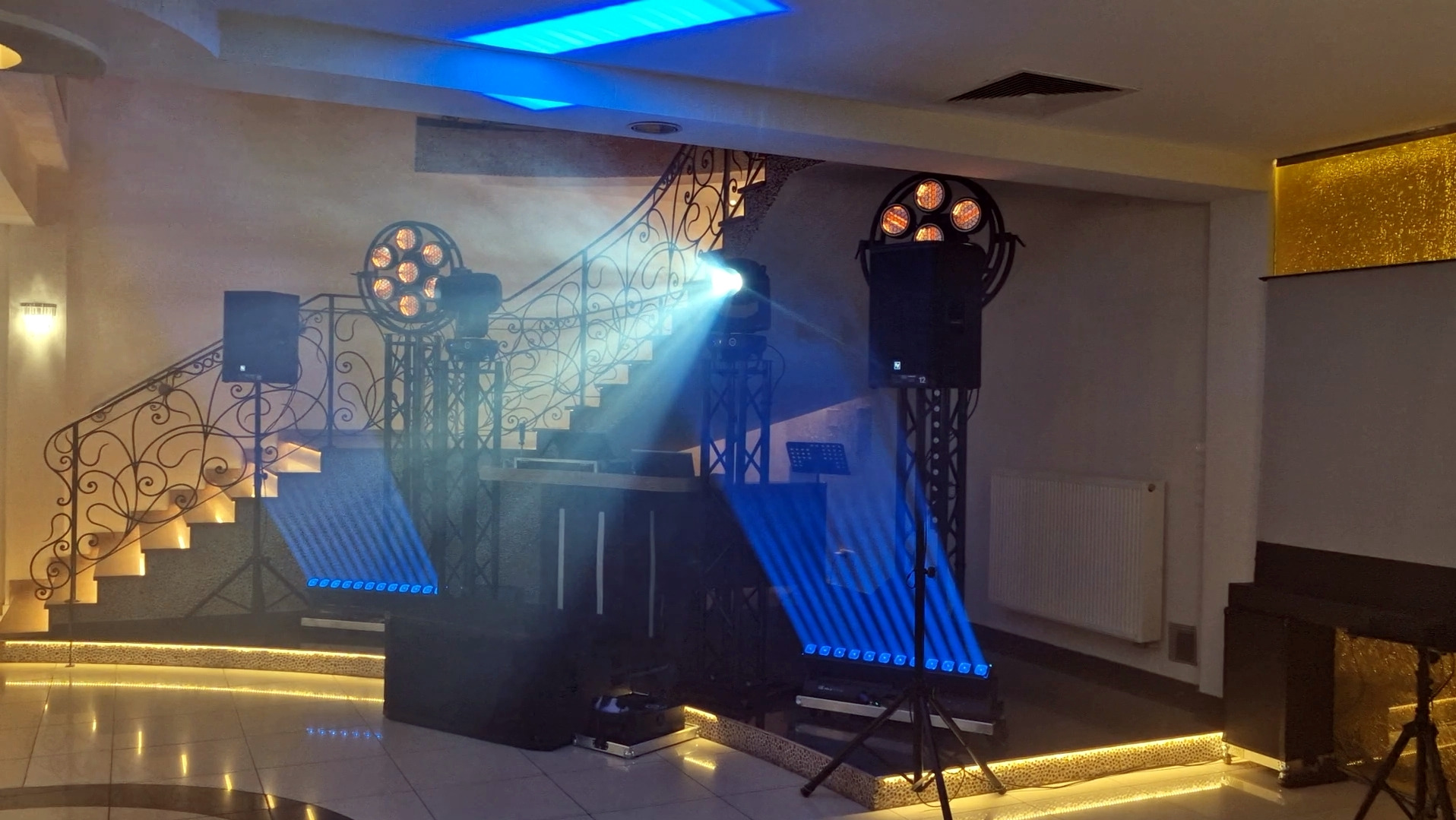 Scena z nagłośnieniem i oświetleniem na tle schodów z ozdobną balustradą. Widoczne reflektory, kolumny głośnikowe i konsoleta DJ-ska. Efekty świetlne w kolorze niebieskim.