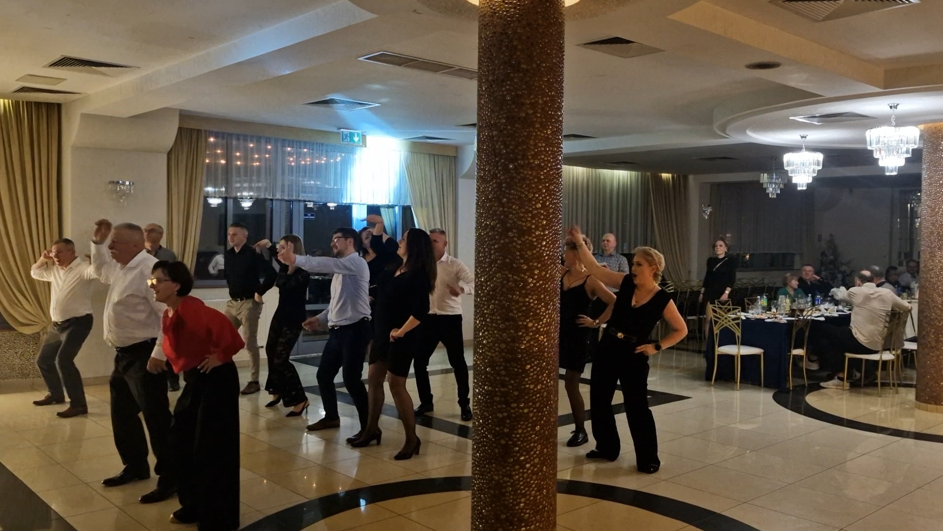Grupa osób tańczy na parkiecie w eleganckiej sali balowej z kolumną. W tle widoczne stoły z gośćmi i dekoracje.