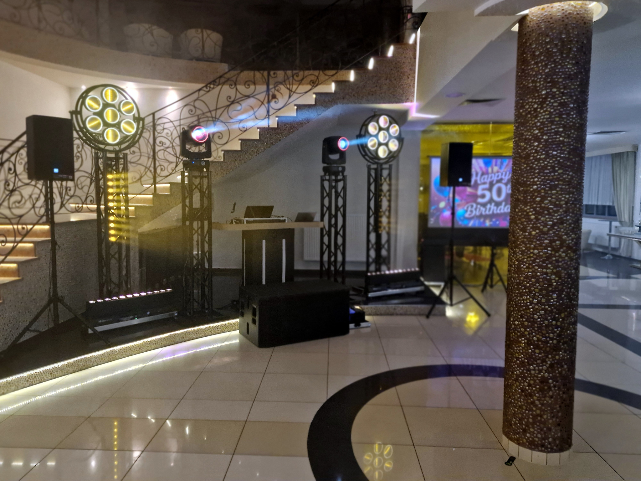 Sala z nagłośnieniem i oświetleniem eventowym, reflektory na statywach, kolumny głośnikowe, schody w tle, dekoracyjny filar. Przygotowania do imprezy.