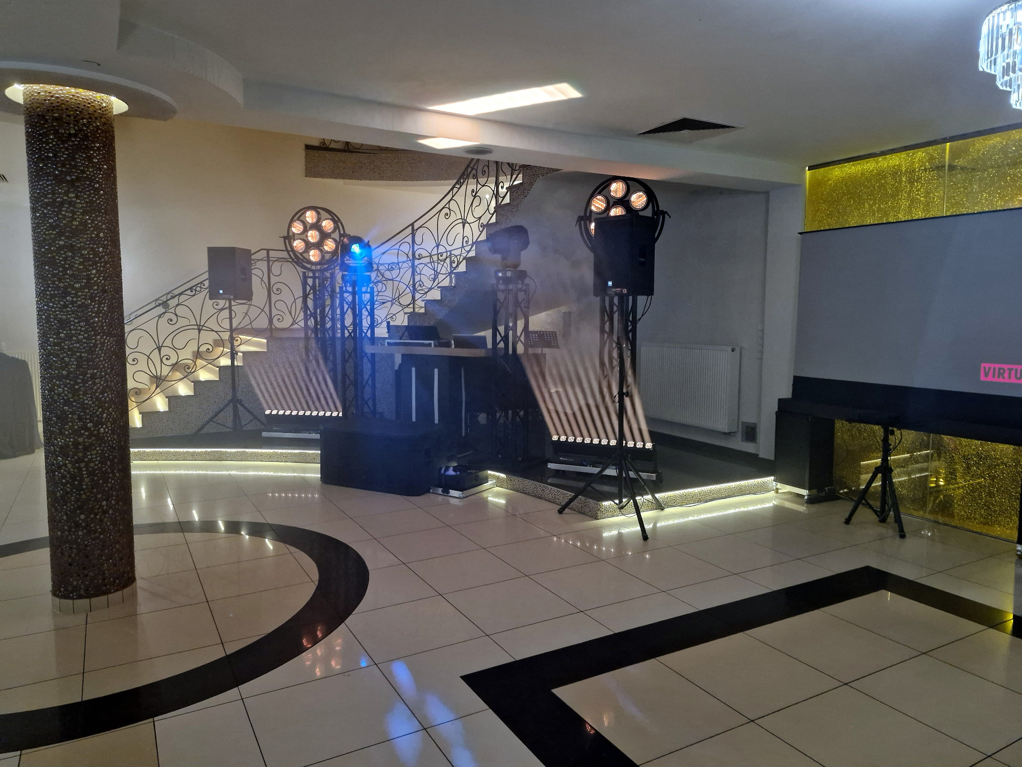 Sala bankietowa z oświetleniem eventowym: reflektory, kolumny głośnikowe, dekoracyjne światła LED przy schodach. Scena gotowa na występ.