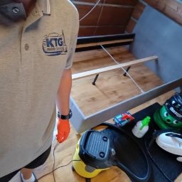 KTG mobile service - Przygotowanie do prania tapicerki łóżka na poddaszu: odkurzacz piorący, środki czystości i osoba w firmowej koszulce z logo KTG Mobile Service.