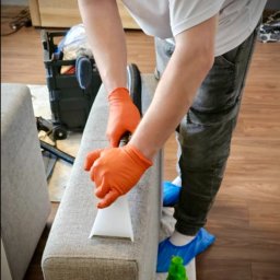 KTG mobile service - Pranie narożnika odkurzaczem ekstrakcyjnym. Osoba w rękawicach i ochraniaczach na butach czyści tapicerkę. W tle sprzęt do czyszczenia i podłoga.