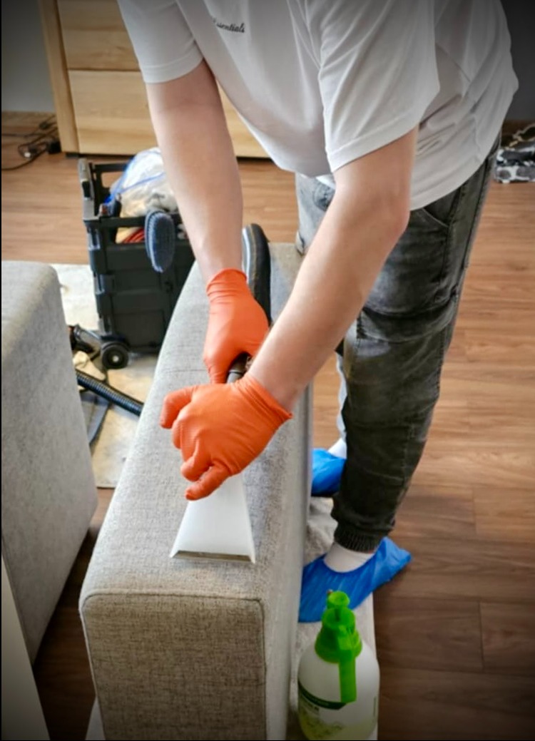 Pranie narożnika odkurzaczem ekstrakcyjnym. Osoba w rękawicach i ochraniaczach na butach czyści tapicerkę. W tle sprzęt do czyszczenia i podłoga.