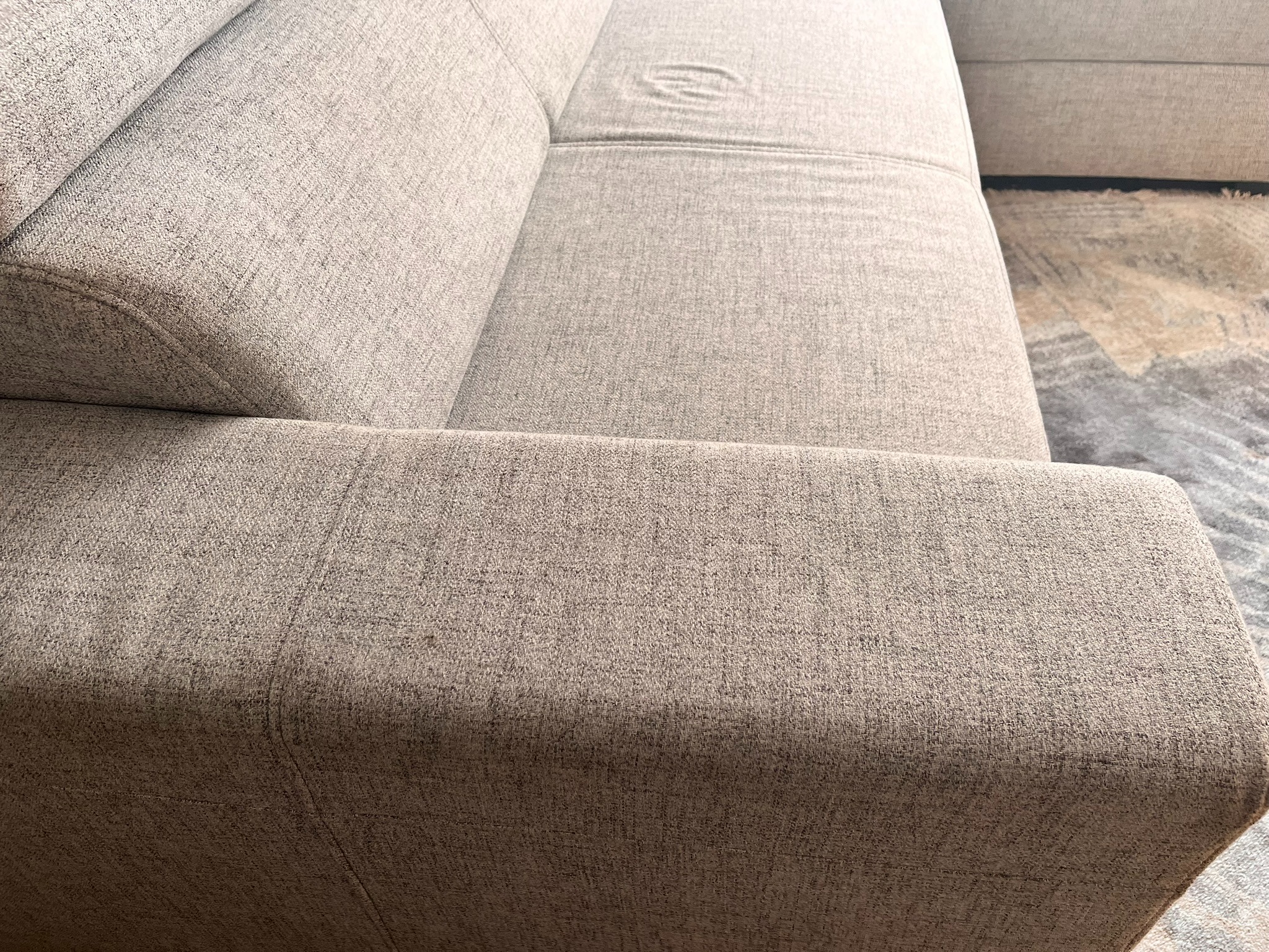 Fragment sofy z jasną tapicerką o wyraźnej fakturze, widoczny szew i delikatne wgniecenie na siedzisku. W tle fragment dywanu.