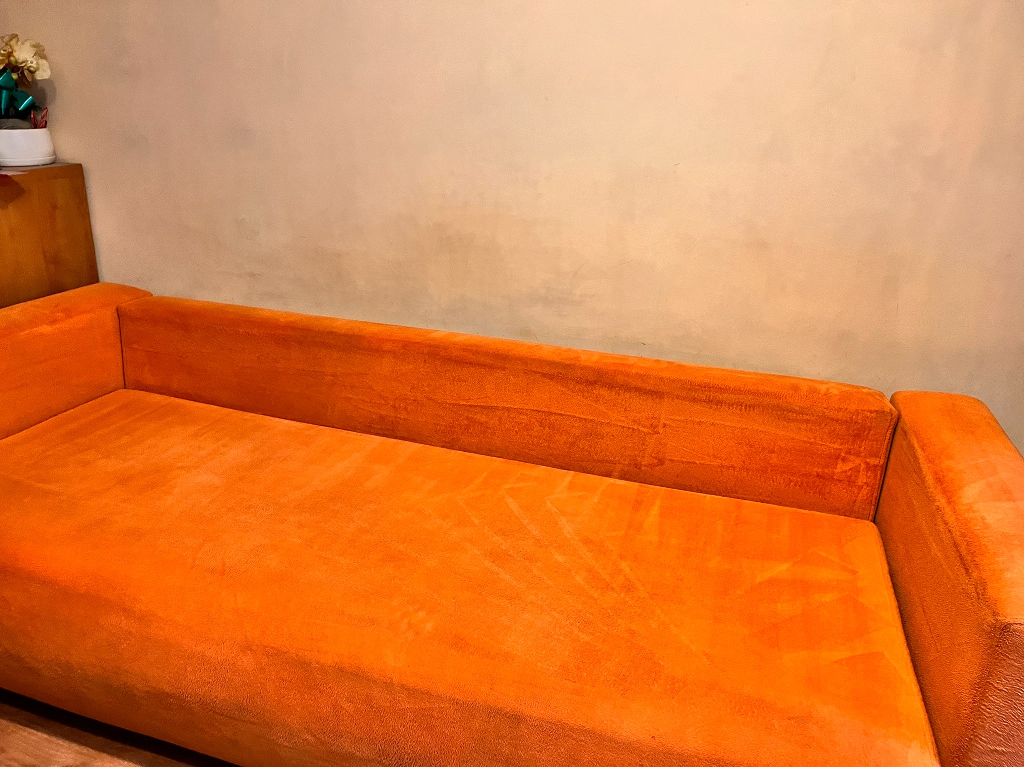 Pomarańczowa kanapa z weluru po czyszczeniu, widoczna faktura materiału i brak plam. Na szafce obok stoi dekoracja roślinna. Ściana w tle w odcieniach beżu.