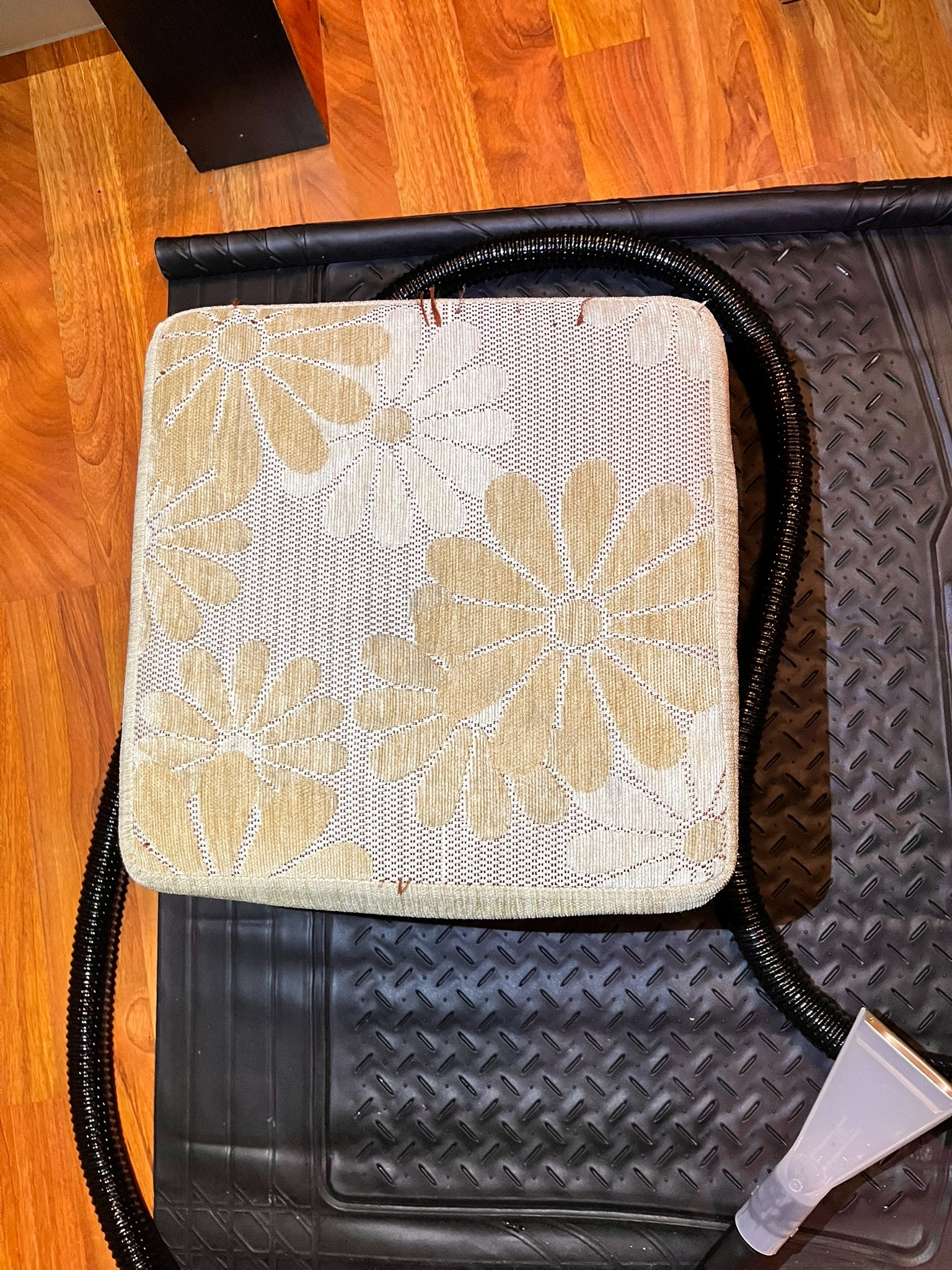 Kwadratowa poduszka z kwiatowym wzorem, umieszczona na czarnej macie, obok węża odkurzacza piorącego. Widoczne drewniane panele podłogowe.