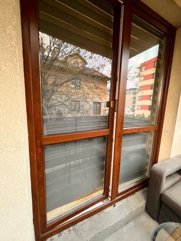 Brązowe, przesuwne okno balkonowe z widokiem na budynki i drzewo. Widoczne ślady po umyciu na szybie. Częściowo widoczny szary fotel balkonowy.