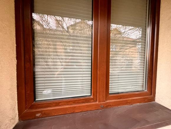 Drewniane okno z żaluzjami w brązowej ramie, widok z bliska. Na szybach odbija się zarys drzew i budynków. Widoczny parapet i fragment elewacji.