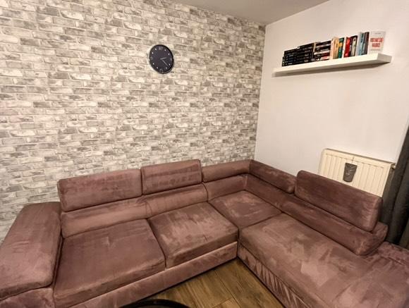 Narożnik z brązowego weluru na tle ściany z cegły i półki z książkami. Widoczne ślady użytkowania na siedzisku.