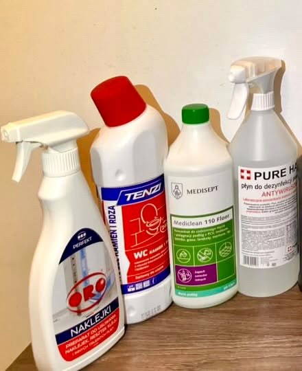Zestaw środków czystości: płyn do WC, podłóg, odplamiacz i dezynfekujący w białych butelkach z kolorowymi etykietami, ustawione na drewnianej powierzchni.