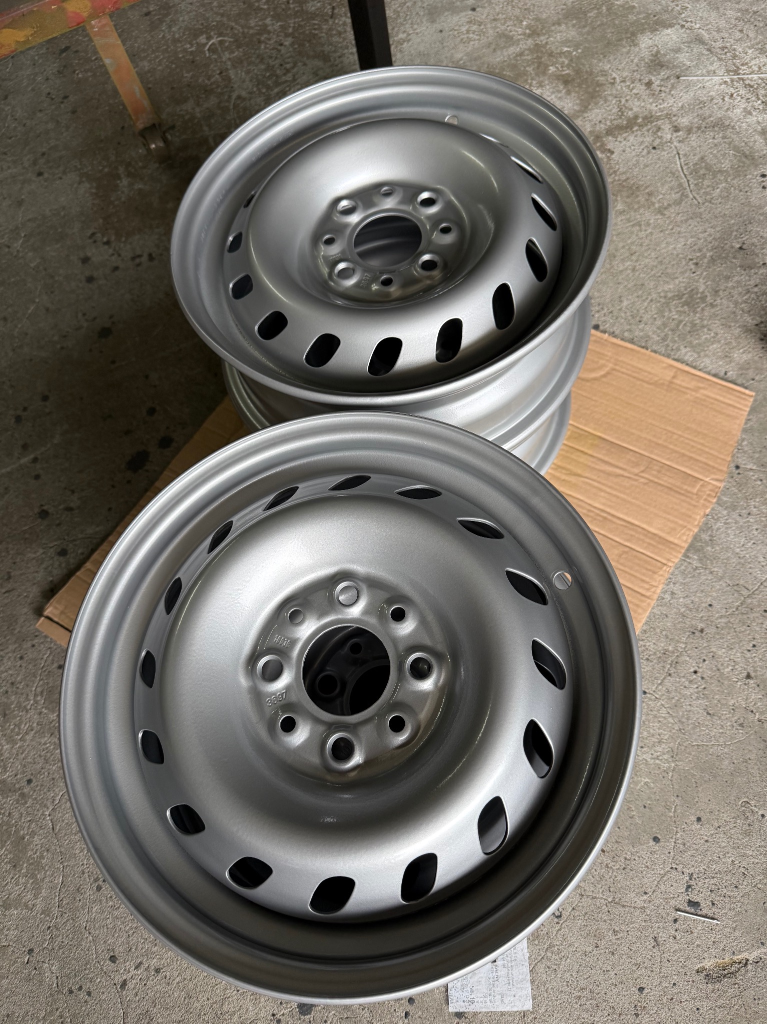 Stos srebrnych felg stalowych na kartonie, po obróbce. Widoczne otwory wentylacyjne i mocujące. Szary beton w tle. Felgi ułożone jedna na drugiej.