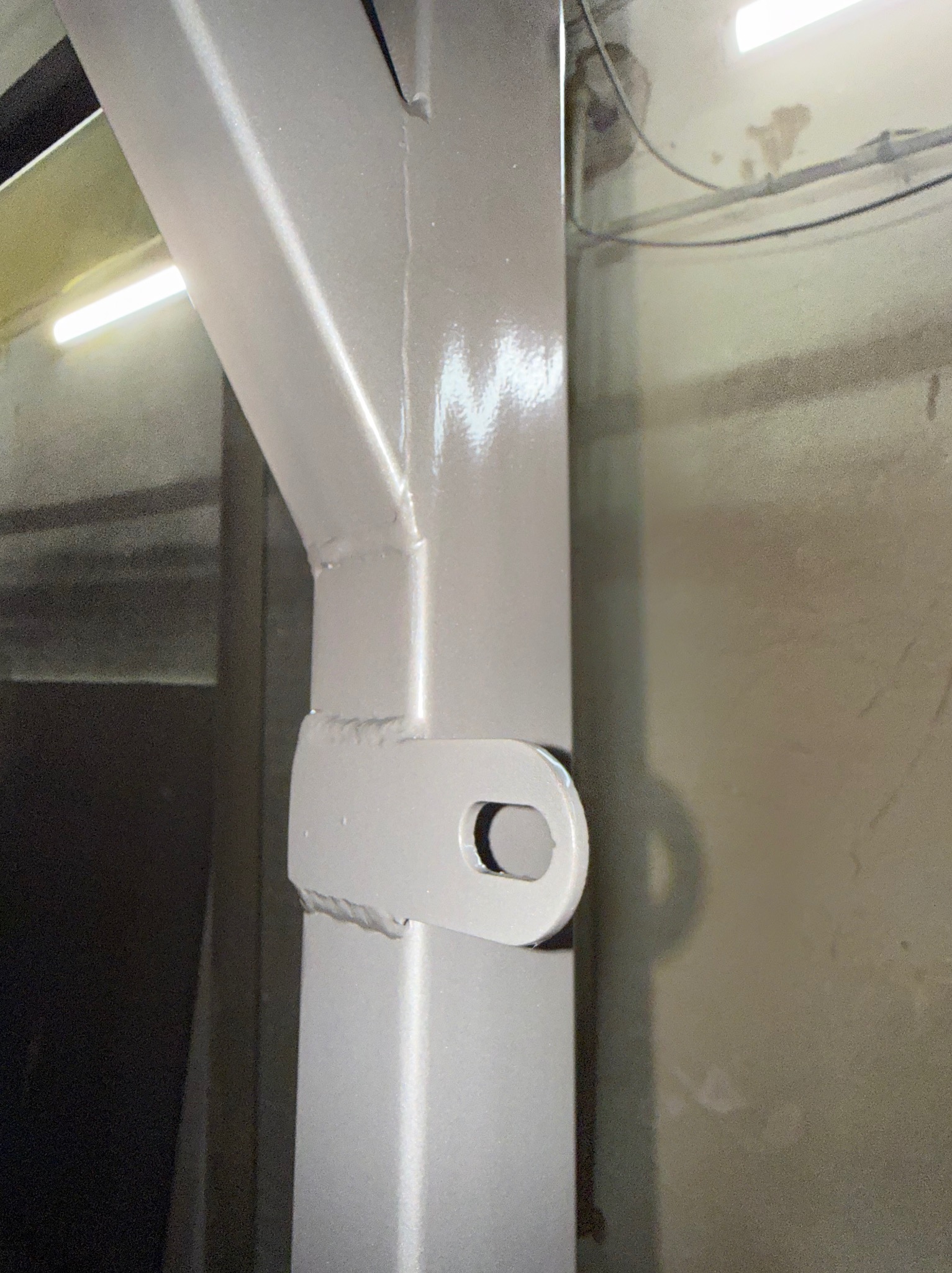 Szara, metalowa konstrukcja z uchwytem, spawana, z widocznymi śladami obróbki. Element konstrukcyjny w surowym wnętrzu, oświetlony sztucznym światłem.