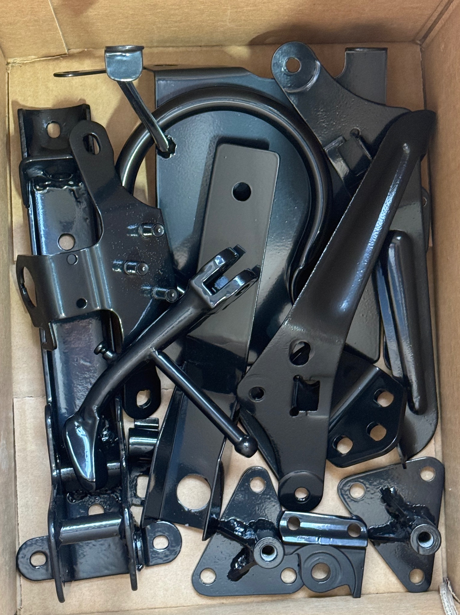 Części metalowe w kartonie, pokryte czarną, błyszczącą powłoką. Elementy o różnych kształtach i rozmiarach, prawdopodobnie po obróbce lub malowaniu, gotowe do montażu.