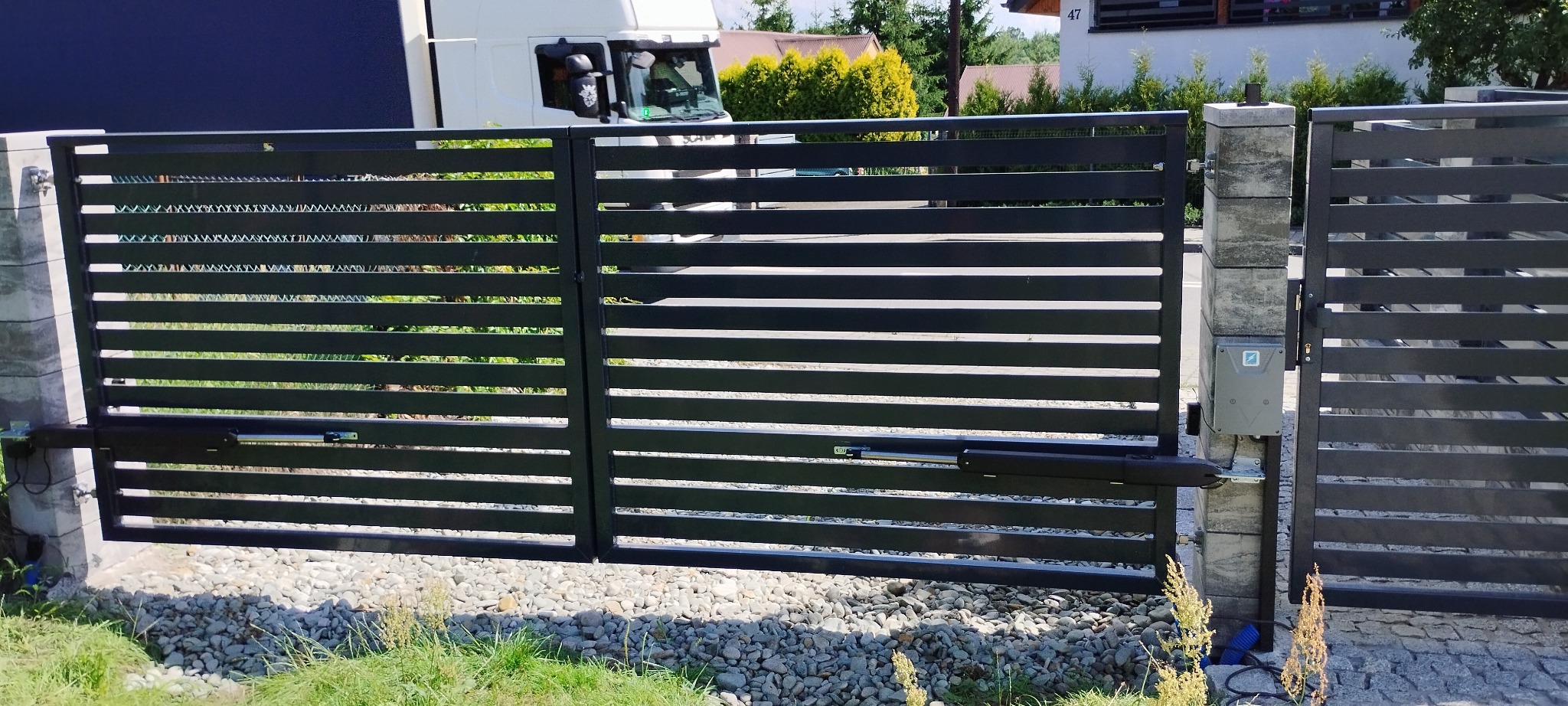 Nowoczesna, pozioma brama przesuwna z automatyką, widoczny napęd i sterownik. Szara kolorystyka bramy i słupków, żwirowe podłoże, zieleń w tle.
