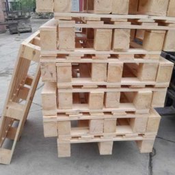 Palety Fitosanitarne I SPRZEDAŻ TARCICY LESZEK MICHALIK - Stos jasnych, drewnianych palet ułożonych jedna na drugiej, obok widoczna kolejna paleta oparta o stos. W tle fragment wózka widłowego i składowane deski.