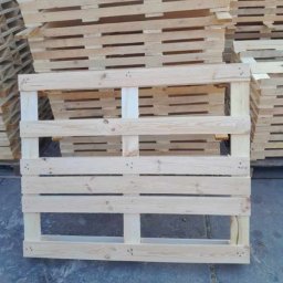 Palety Fitosanitarne I SPRZEDAŻ TARCICY LESZEK MICHALIK - Jasna, drewniana paleta euro na tle stosu palet. Widoczne słoje drewna i metalowe zszywki. Paleta ustawiona na szarym podłożu.