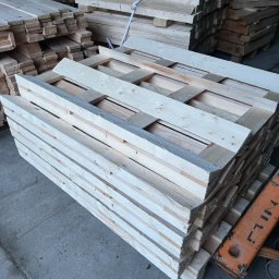 Palety Fitosanitarne I SPRZEDAŻ TARCICY LESZEK MICHALIK - Jasne, nowe palety drewniane ułożone w stos w magazynie w Zambrzyńcu. Widoczne deski i konstrukcja palet. W tle inne stosy palet i fragment wózka widłowego.