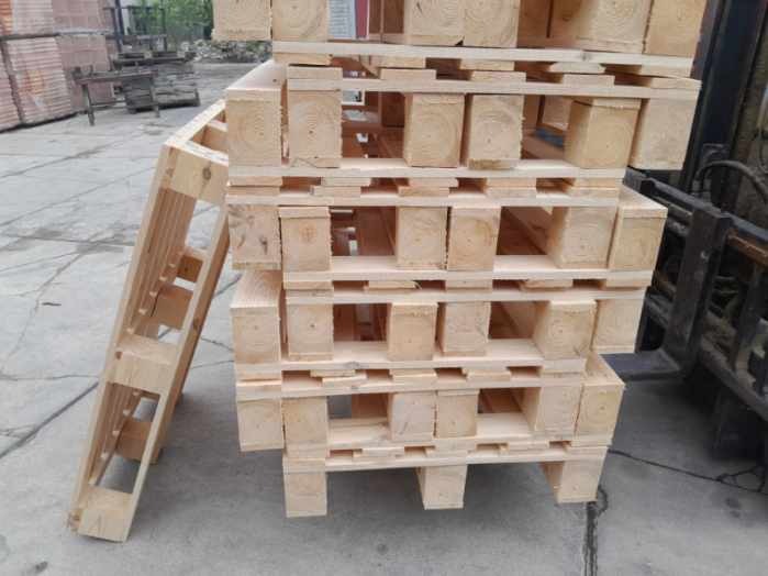 Stos jasnych, drewnianych palet ułożonych jedna na drugiej, obok widoczna kolejna paleta oparta o stos. W tle fragment wózka widłowego i składowane deski.