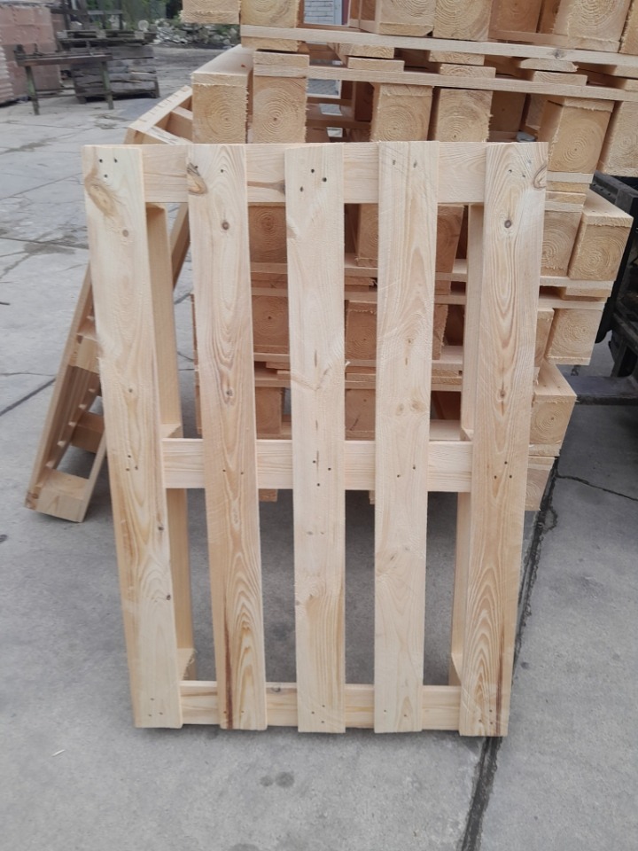 Jasna, drewniana paleta na szarym tle, z widocznymi słojami drewna i konstrukcją. W tle stos palet, tworzący wzór powtarzalnych elementów.