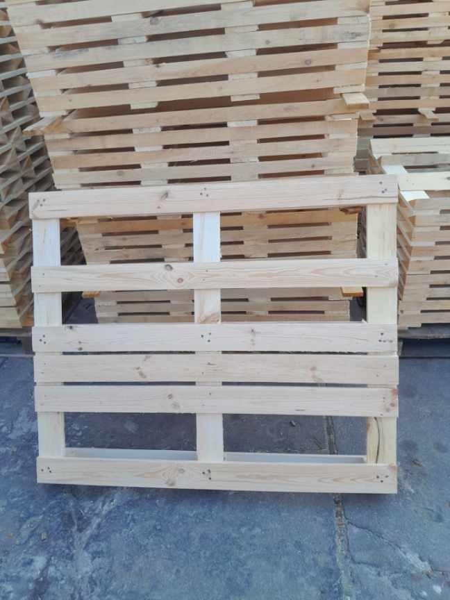 Jasna, drewniana paleta euro na tle stosu palet. Widoczne słoje drewna i metalowe zszywki. Paleta ustawiona na szarym podłożu.