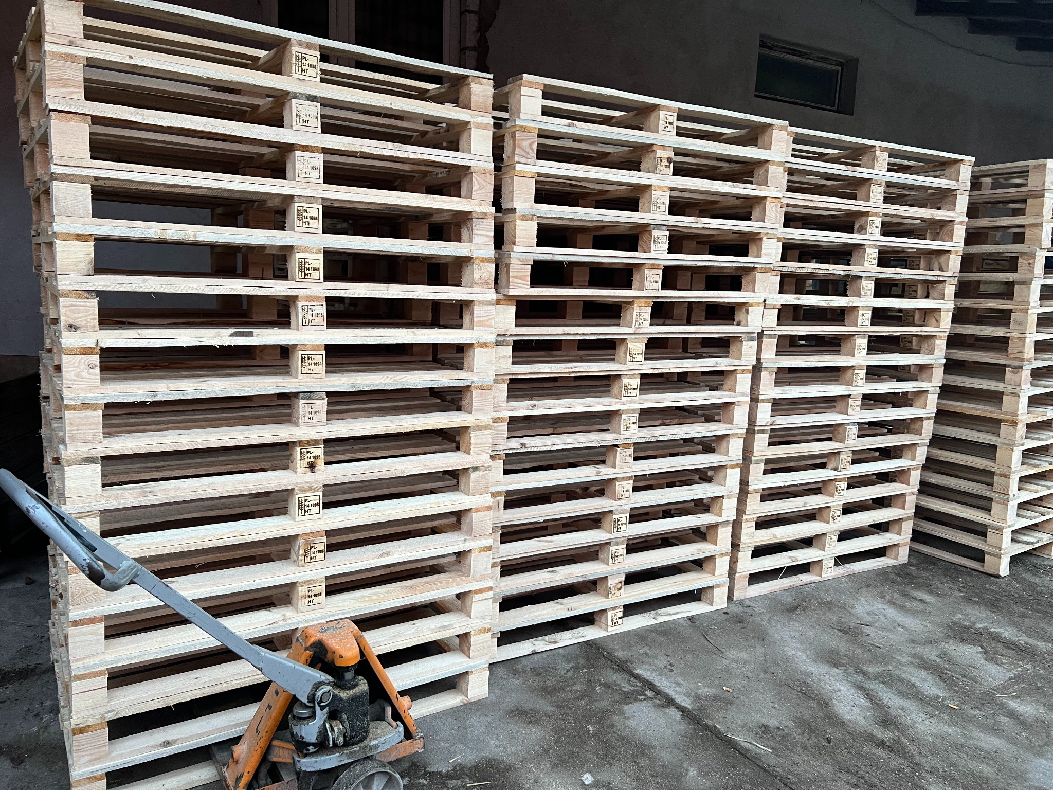 Stosy jasnych, drewnianych palet euro ułożone w magazynie, z wózkiem paletowym z przodu. Widoczne oznaczenia na paletach. Surowe, betonowe tło.