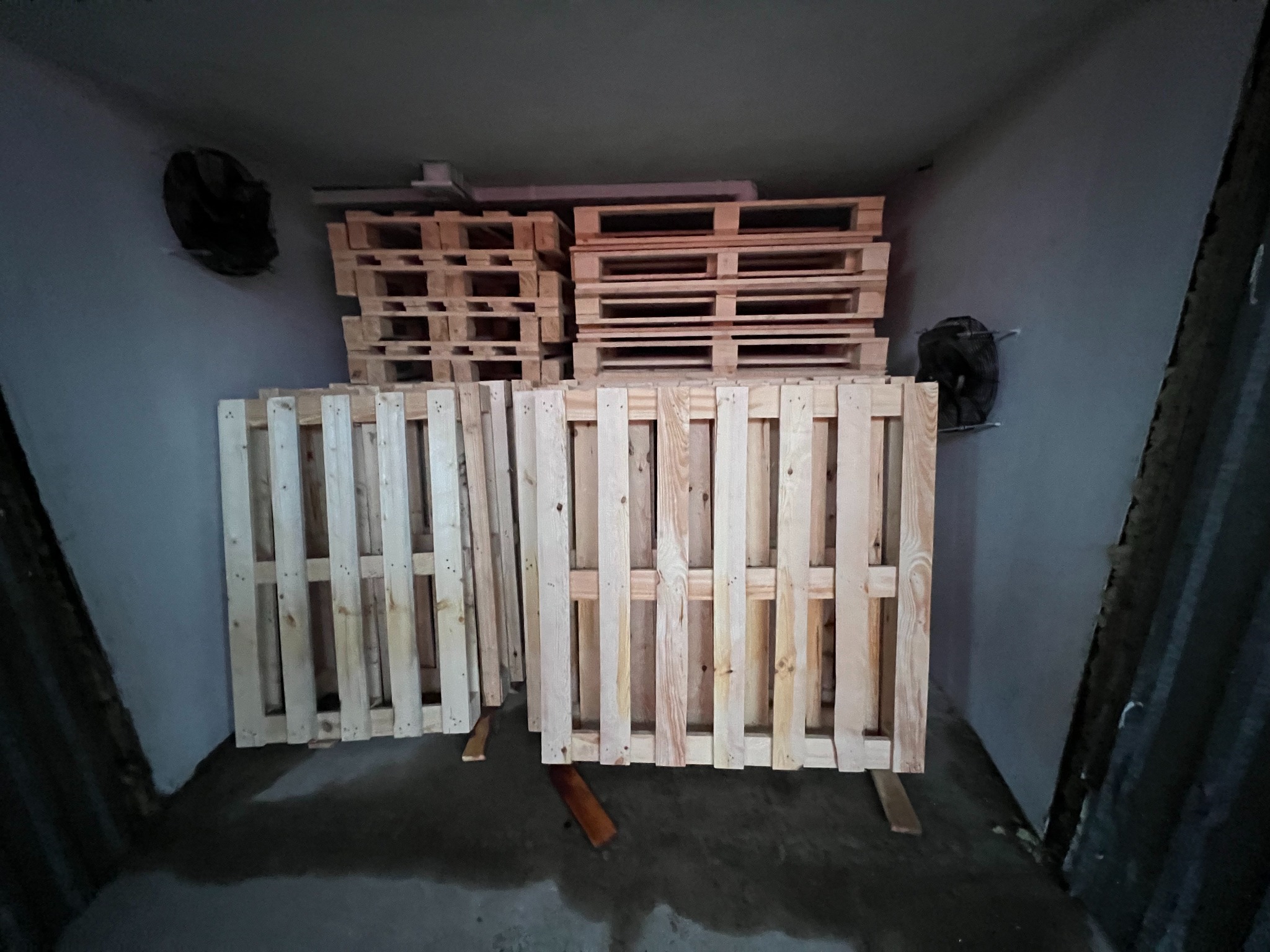 Stos drewnianych palet w magazynie z betonową podłogą i wentylatorami na ścianach. Palety ułożone w stosy, widoczne detale konstrukcji i faktura drewna.