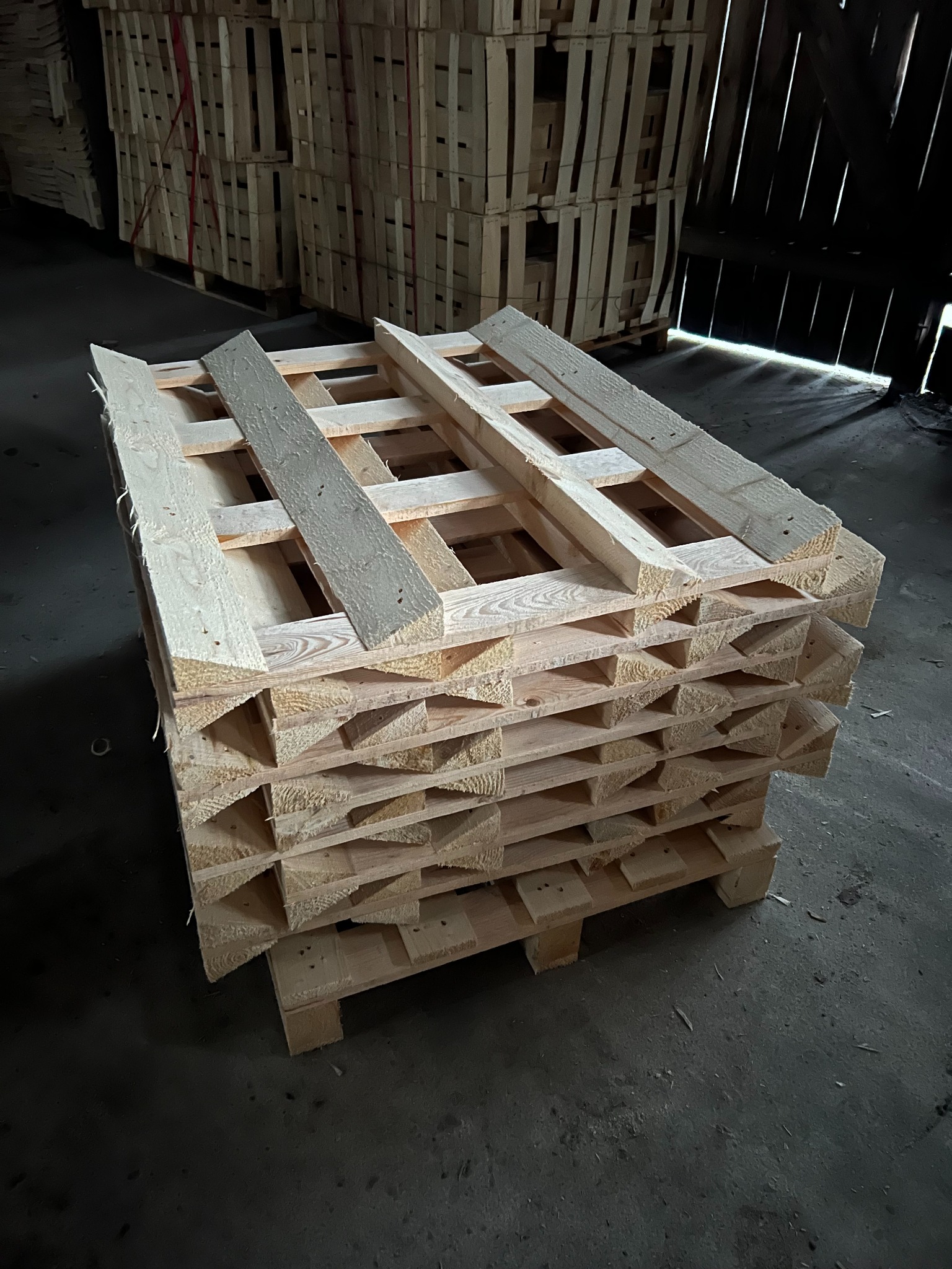 Stos jasnych, drewnianych palet w magazynie, ułożonych jeden na drugim. W tle widoczne inne palety i ciemne ściany magazynu. Surowe drewno, widoczne słoje.