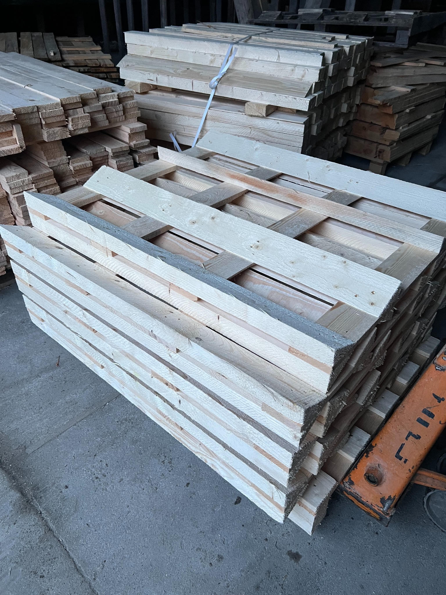 Jasne, nowe palety drewniane ułożone w stos w magazynie w Zambrzyńcu. Widoczne deski i konstrukcja palet. W tle inne stosy palet i fragment wózka widłowego.